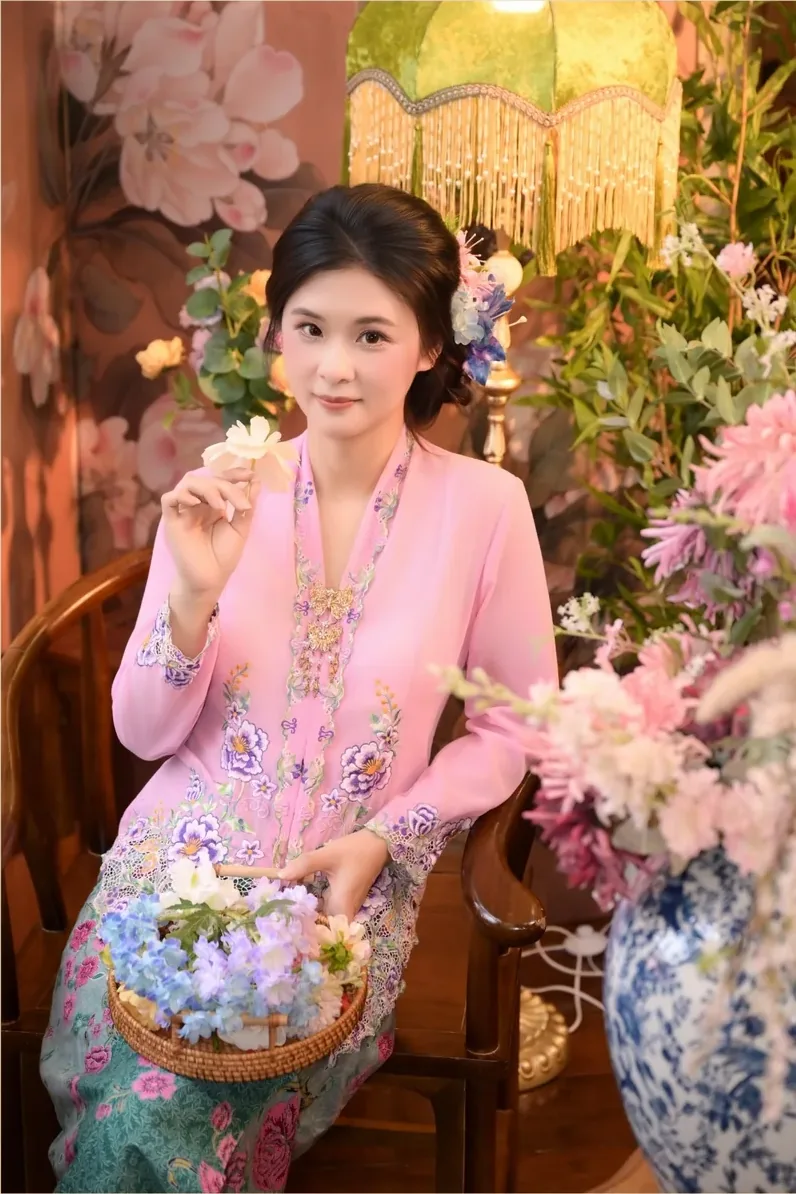 身穿粉色绣花娘惹衫的女子手捧花篮，在摄影棚布景前